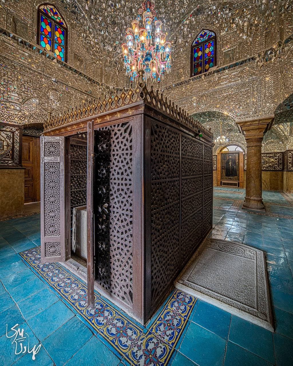 ضریح چوبی مشبک اطراف مقبره در تالاری با کف‌پوش کاشی فیروزه‌ای و سقف آینه‌کاری